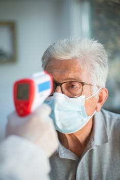 A senior man with white hair and eyeglasses gets his temperature checked using a digital thermometer.